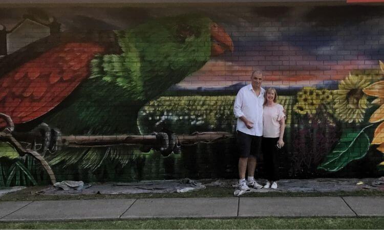 A man and a woman standing in front of a colorful mural of a parrot.