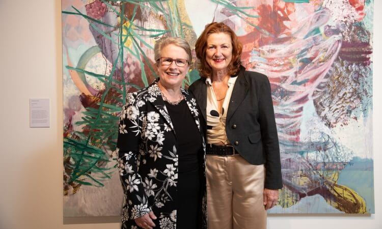 Two women smiling in front of a colorful abstract painting in an art gallery.