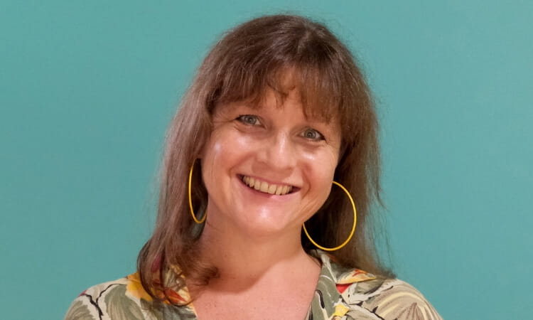 Woman with a friendly smile, wearing hoop earrings and a floral top against a turquoise background.