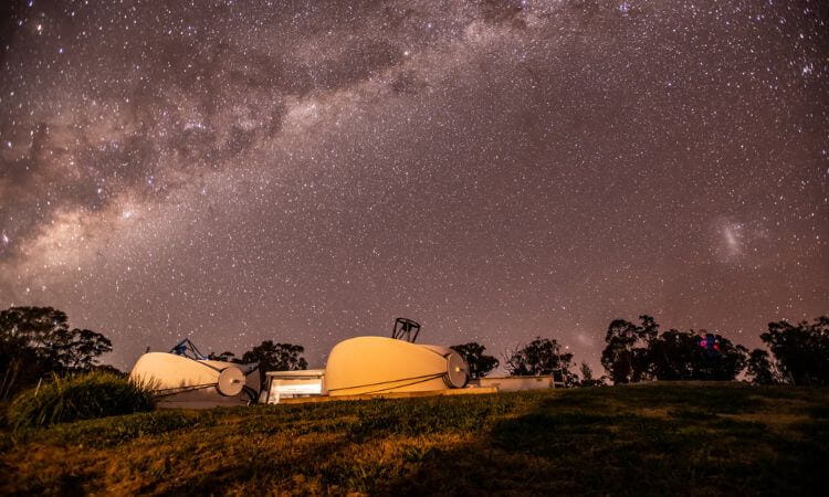 Astronomical observatory at night under a starry sky, featuring multiple telescope domes open against a backdrop of the milky way.