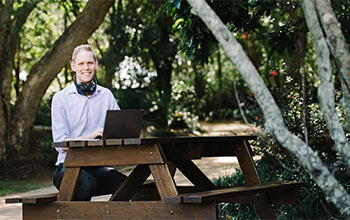 Dan is sitting at on outdoor bench with his laptop, smiling at the camera.