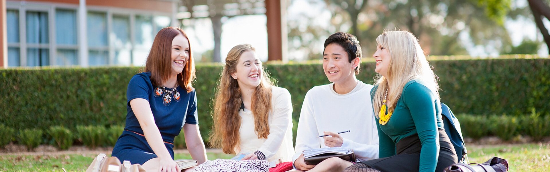 Four students sitting outdoors on the grass, sharing a conversation with textbooks and notebooks.