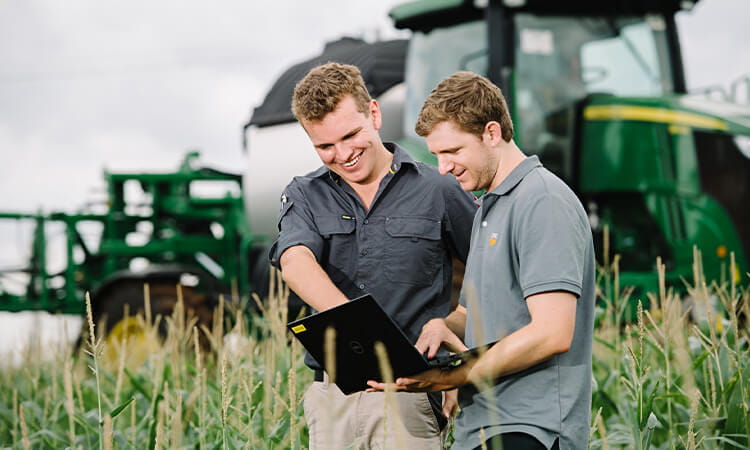 Two man standing in a field, in front of a tractor, looking at a laptop. 